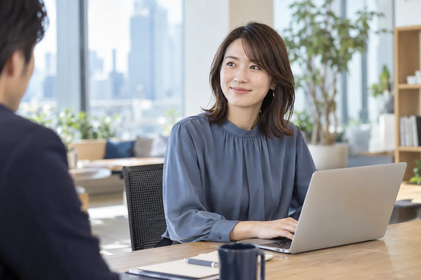 明るいオフィスで、同僚と話しながらパソコンに向かう女性。AI生成の就業イメージ。
