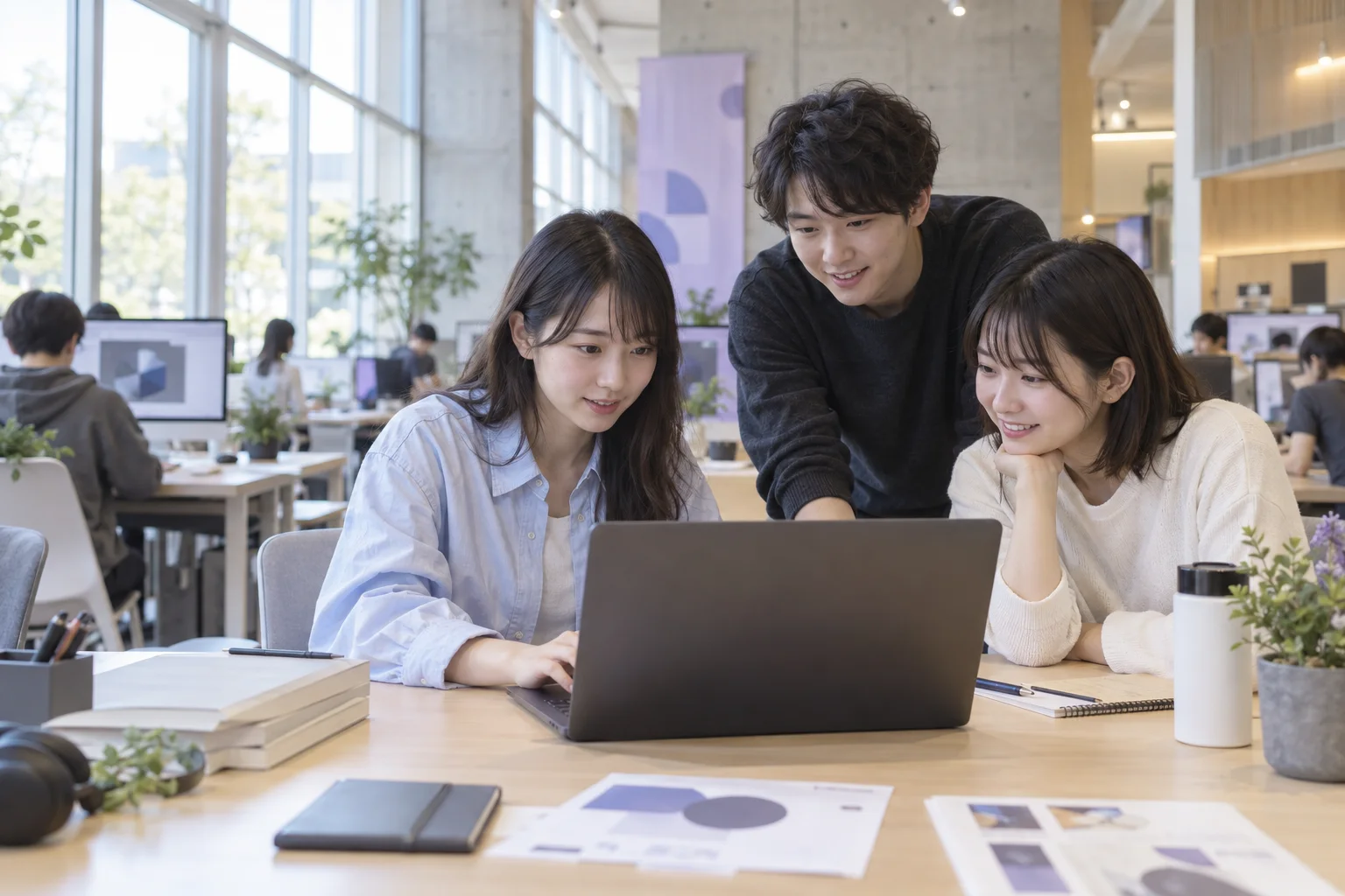 明るいコンピューターラボで、3人の学生が同じ画面を見ながら制作を楽しむ架空のキャンパス風景。